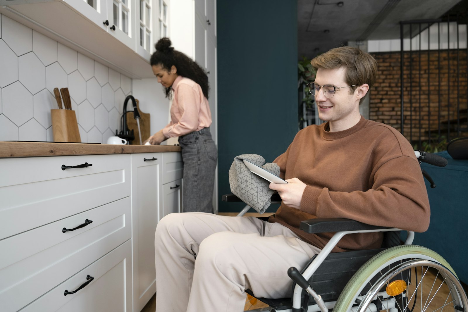 An NDIS participant using a tablet at home while a support worker works nearby in the kitchen